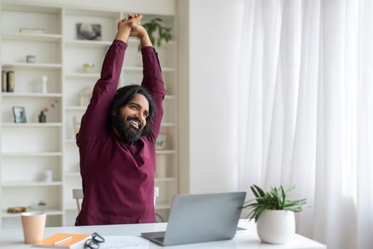Happy indian man stretching his arms after working on laptop at home