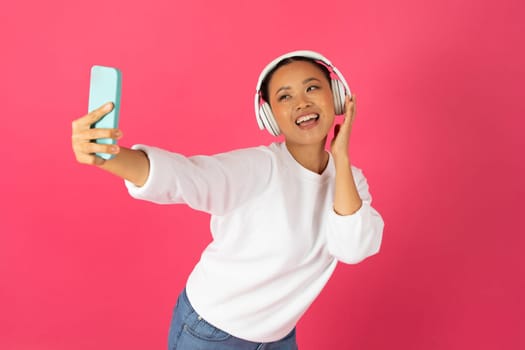 Joyful asian woman wearing wireless headphones talking selfie on smartphone