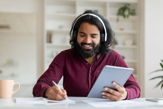 Indian man enjoying music on headphones while browsing tablet and taking notes