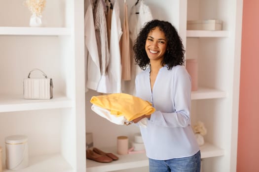 Cheerful black woman holding folded yellow shirt, wardrobe behind