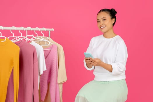 Smiling Asian Woman Using Her Mobile Phone While Sitting Near Clothing Rack