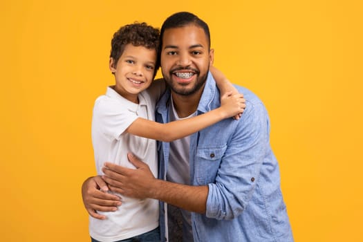 Affectionate black father and little son embracing with joyful smiles, happy african american dad and male kid wearing casual clothes posing together against yellow studio background, free space