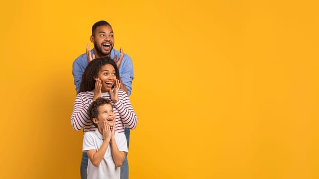 Amazed black family with little son looking aside at copy space against yellow studio background, cheerful parents and male kid expressing curiosity and surprise, emotionally reacting to nice offer