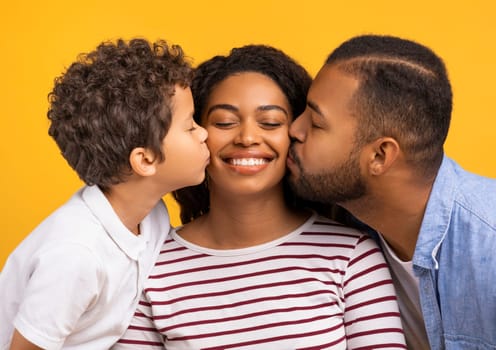 Adorable son and loving husband giving kisses on cheeks of smiling black mother, showing love and affection, greeting their mom and wife with birthday or womens day, standing on yellow background