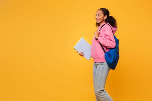 Cheerful black teenager student girl carrying blue backpack, yellow studio