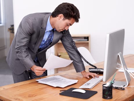 Businessman, phone call and multitasking on computer in office, networking and technology in workplace. Young accountant, talking or landline with paperwork, consultation or finance advice in company
