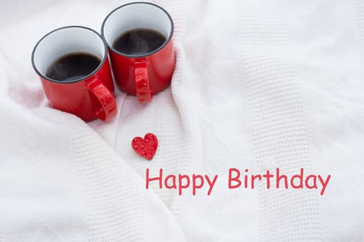 Romantic still life, two red cups of coffee on a white plaid. Happy birthday lettering, holiday card