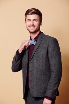 Portrait, fashion and smile with a young man in studio on a tan background to wear a trendy bowtie. Happy, style and clothes with a confident model in a retro or vintage suit for a formal event