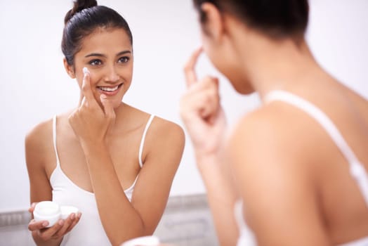 Woman, skincare and moisturizing in bathroom mirror, lotion and face cream on skin for hydration. Happy female person, reflection and cosmetics or dermatology, self care and apply sunblock at home