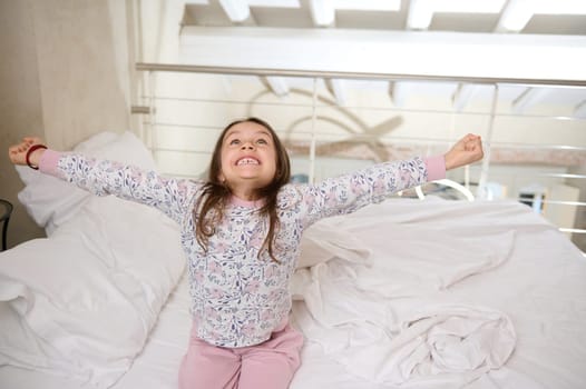 Authentic cheerful mischievous little girl waking up in the morning, stretching her arms upwards