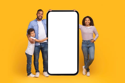 Happy Black Parents And Their Little Son Standing Near Big Blank Smartphone