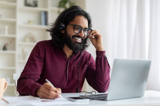Young indian man wearing headset using laptop and smiling while taking notes