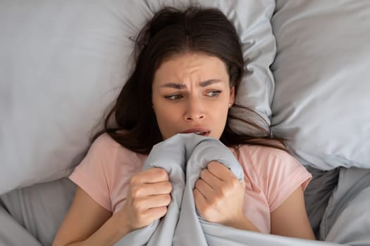 Scared young woman lying in bed be afraid of sounds or unable to get to sleep and pulling blanket to face, top view