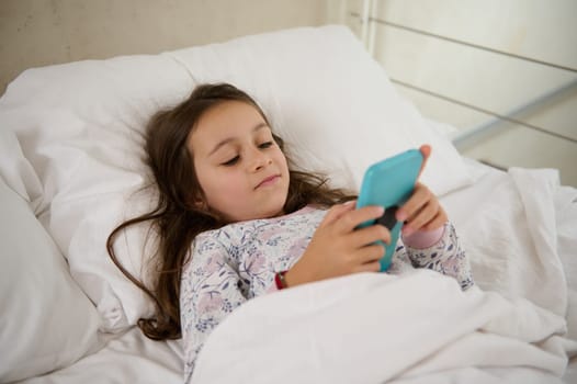 Cute toddler girl playing with a smartphone, lying down in a comfortable double bed while wake in up in the morning