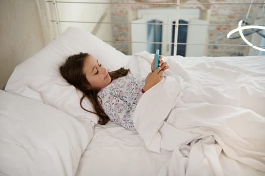 Adorable elementary age kid, little child girl using smartphone, watching movies while lying on the bed in the morning.