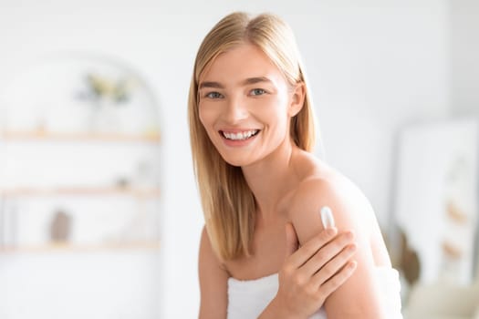 blonde lady in white towel applies moisturizer on shoulder indoor