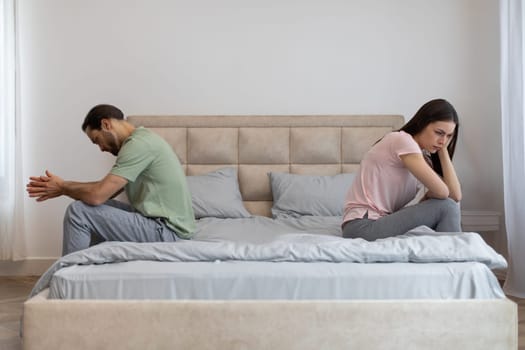 Young couple having relationship difficulties, sitting on opposite sides of bed, ignoring each other at home