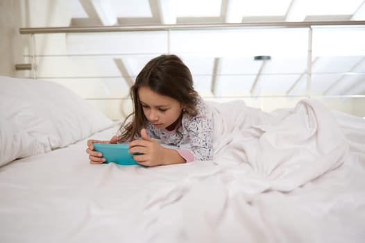 Internet addiction in childhood. Little girl having fun on smartphone while lying on bed. The concept of digital gadgets