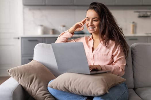 Happy young woman browsing on laptop while comfortably lounging on sofa