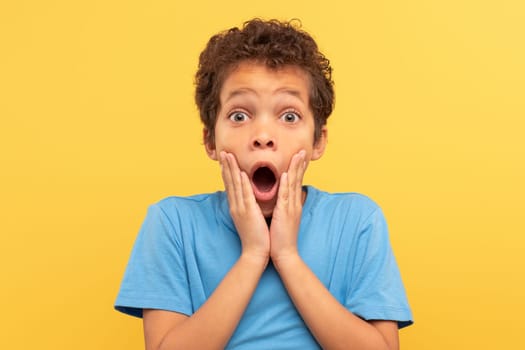 Shocked boy with hands on face, open mouth on yellow background