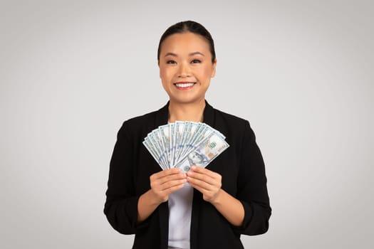 A radiant Asian businesswoman in professional attire beams as she offers a fan of hundred-dollar bills