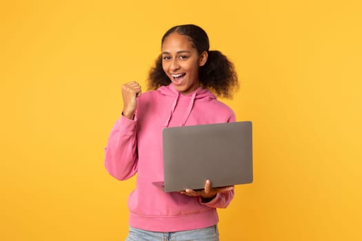 Excited black teenager girl with laptop making YES gesture, studio