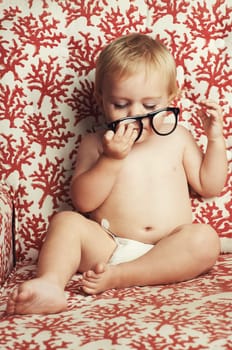 Glasses, playing and sweet baby in a studio with vision, health or eye care accessory for development. Cute, eyewear and young child, infant or toddler kid with spectacles by wallpaper background.