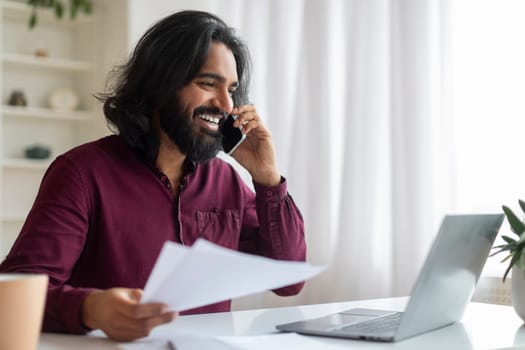 Joyful indian man making phone call, holding papers and using laptop