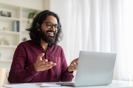 Enthusiastic indian man wearing glasses using headset during online call on laptop