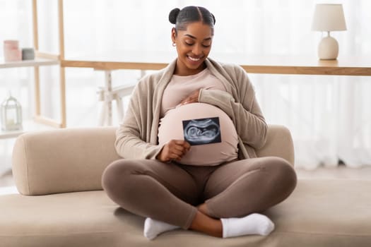 Happy black pregnant woman sitting on couch with her baby sonography photo, smiling african american expectant lady enjoying pregnancy and maternity, showing first image of her child, copy space
