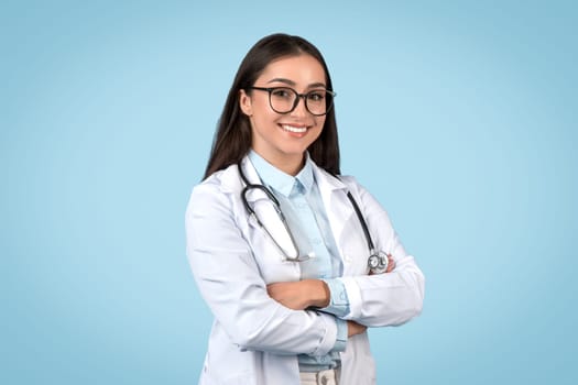 Female professional general practitioner in glasses posing with folded arms on blue studio background