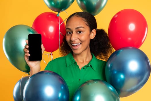 Happy black teenager girl poses with cellphone among balloons, studio