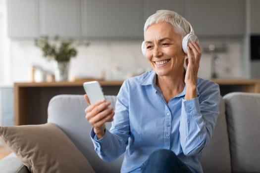 Relaxed mature woman listening music on smartphone with headphones indoor