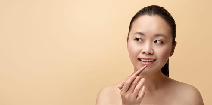 Cheerful millennial chinese naked woman applies pencil to lips and makes basic nude makeup, isolated on beige background, studio, panorama, close up. Natural beauty care, cosmetics