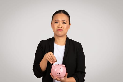 Worried Asian businesswoman in a black blazer deposits a coin into a piggy bank