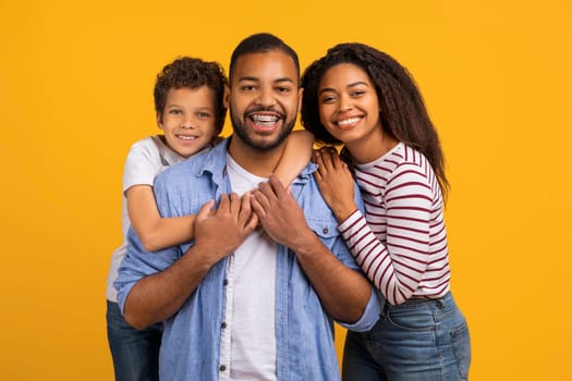 Happy black family of three embracing while posing together in studio, cheerful african american parents and little son sharing affectionate hug, smiling at camera, standing against yellow background