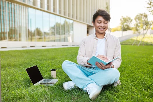 Asian guy student attending webinar, sitting on lawn