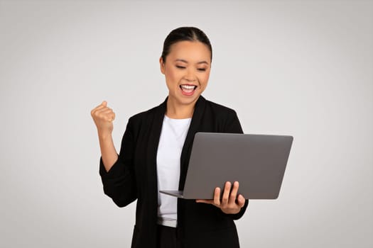 Exuberant Asian businesswoman celebrating success with a fist pump while holding a laptop