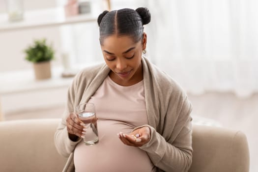 Healthy young pregnant african american woman holding pills, glass of water, expecting lady take daily medicine vitamins supplements for health care during pregnancy, sit on couch at home, copy space