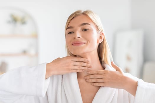 Pretty blonde woman applying moisturizing cream on her neck indoors