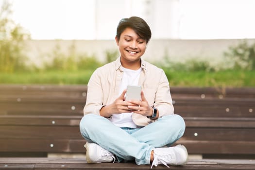 Smiling young chinese guy using smartphone, chilling at park