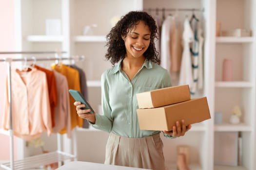 Cheerful young business owner with measuring tape around neck holding cardboard boxes and smartphone, managing orders in her trendy clothing shop, with colorful garments on rack in background