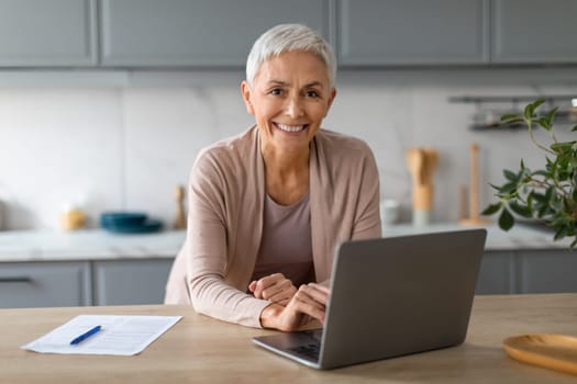Cheerful mature lady typing on laptop smiling to camera indoors