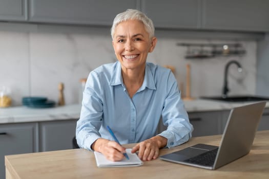 Happy senior businesswoman sitting at laptop writing papers at kitchen