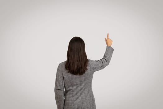 Back view of businesswoman pointing on virtual screen, touching invisible button, grey studio background