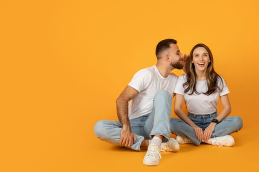 A joyful young couple in casual clothing sits cross-legged on a vibrant orange background