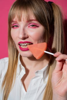 Portrait of a young woman with braces and bright makeup eating a lollipop on a pink background. Vertical photo