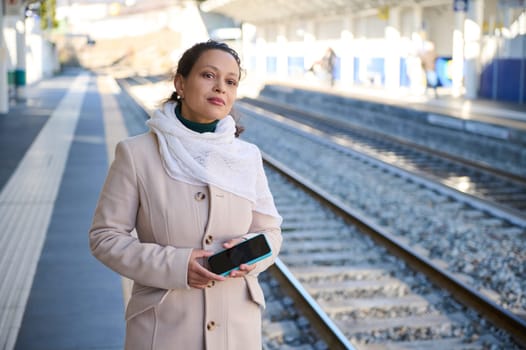Young woman commuter traveling, standing with a smartphone in her hands in a train station, looking into the distance