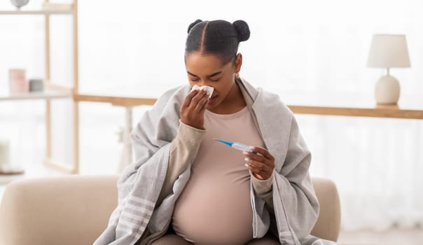 Sick pregnant black woman sneezing and checking body temperature