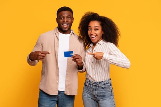 Joyful black couple pointing at credit card on yellow background